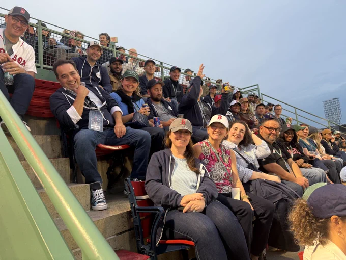 Group of diverse fans smiling and enjoying a baseball game in stadium seats under a cloudy sky.