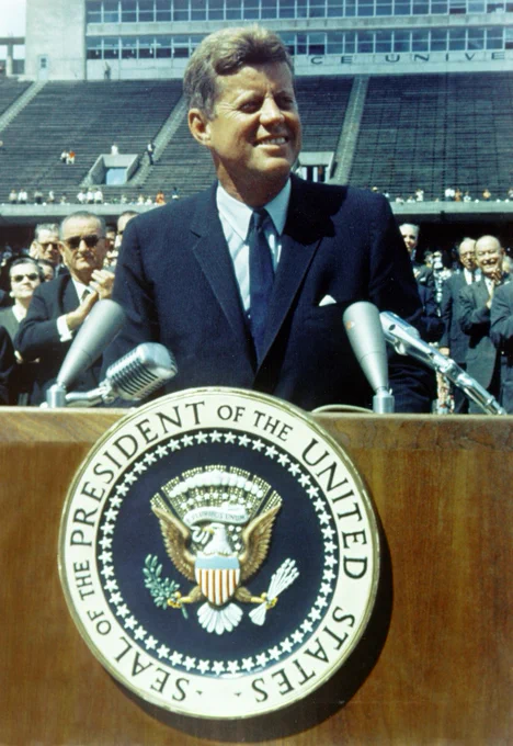 President John F. Kennedy smiles while standing behind a wooden podium bearing the presidential seal during his 1962 speech at Rice University in Houston. Several microphones are positioned in front of him, while people behind him applaud and the stadium stands rise in the background.