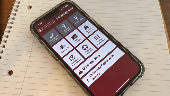 A smartphone displays the UChicago Safe app on a lined notebook. The app's main screen shows menu options like Emergency Contacts, Mobile BlueLight, Friend Walk, Report a Tip, Safety Toolbox, Getting Around, Emergency Procedures, Support Resources, About/Preferences, UChicago Help, University Community, Safety.