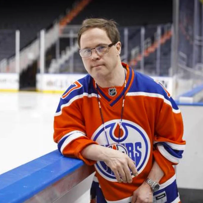 Joey Moss in an orange Edmonton Oilers hockey jersey leans on the boards at an ice rink.