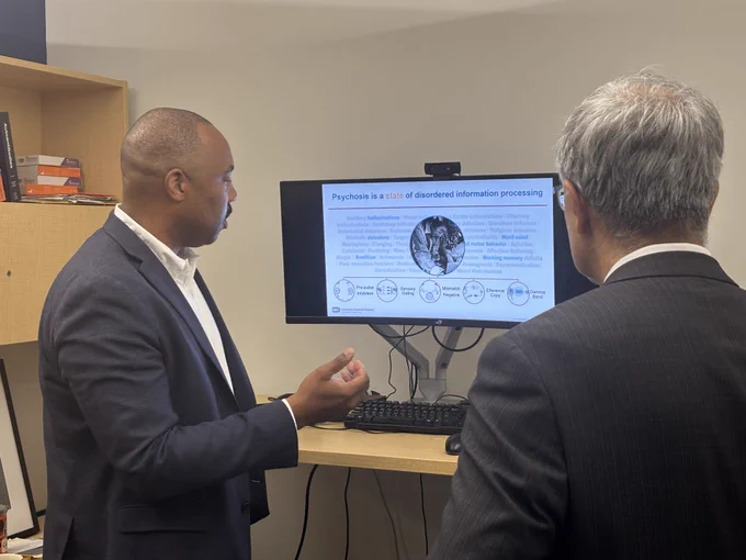 Dr. Jay Bhattacharya looks on at a screen with a slide reading "Psychosis is a state of disordered information processing". An NIMH researcher explains the slide.